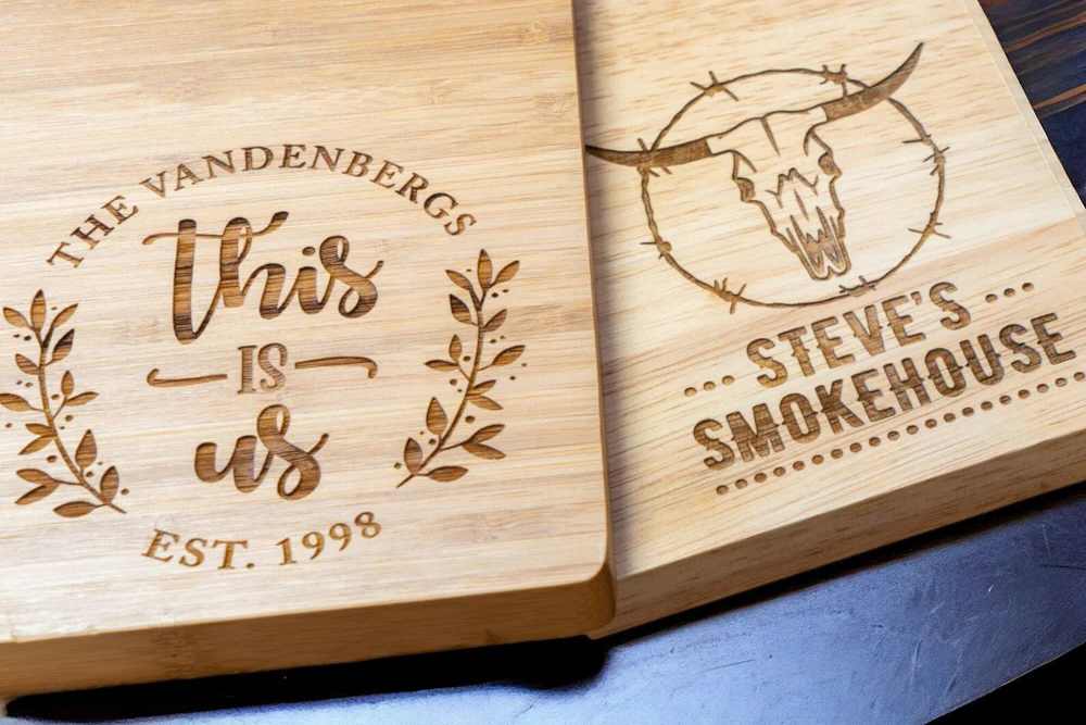 Personalized wooden cutting boards feature intricate engravings: one with "The Vandenbergs - This Is Us - Est. 1998," and the other, "Steve's Smokehouse" with a bull skull design.