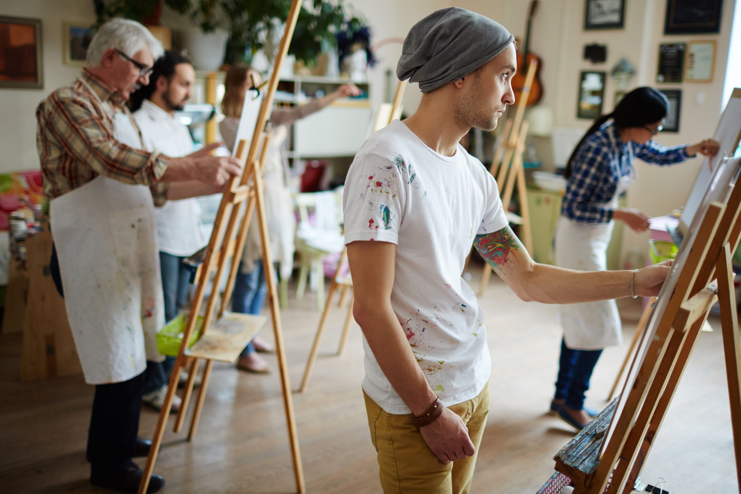 Artists at work in a vibrant studio; a young man focuses on painting while others engage on their easels, showcasing a creative atmosphere.